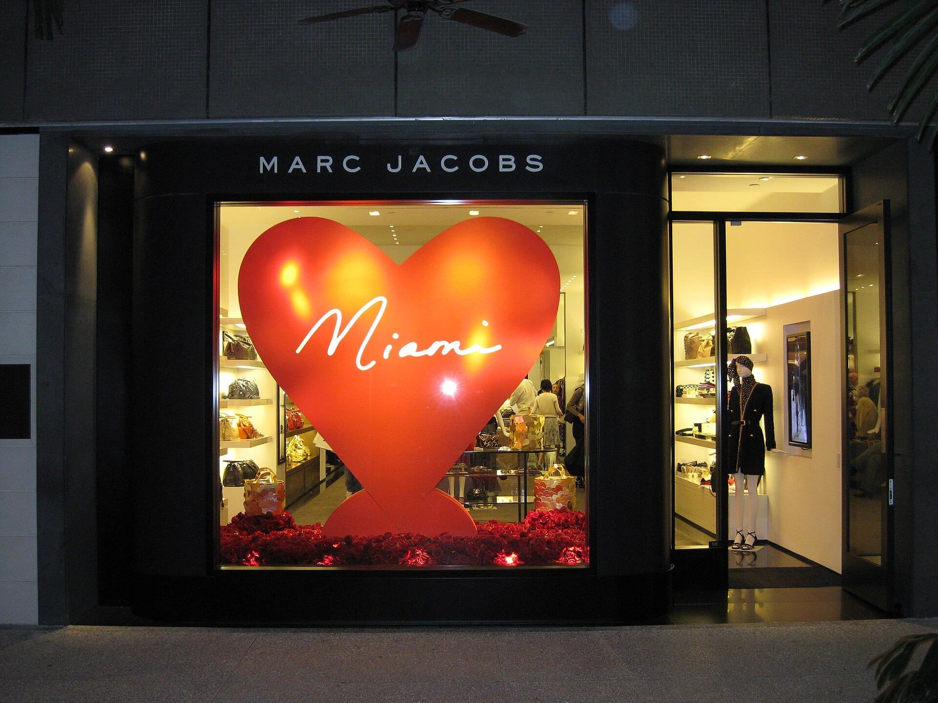 La vetrina del negozio Marc Jacobs al Bal Harbour Shops presenta un grande cuore rosso con 'Miami' scritto all'interno, illuminato di notte con esposizioni eleganti.