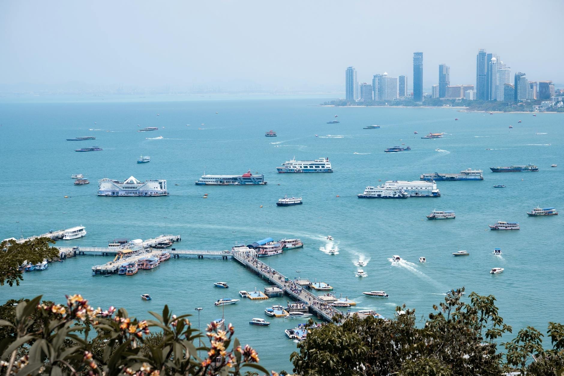 Vista panoramica del Molo Bali Hai a Pattaya con numerosi traghetti e barche attraccati, mare turchese, fiori in primo piano e lo skyline della città sullo sfondo.