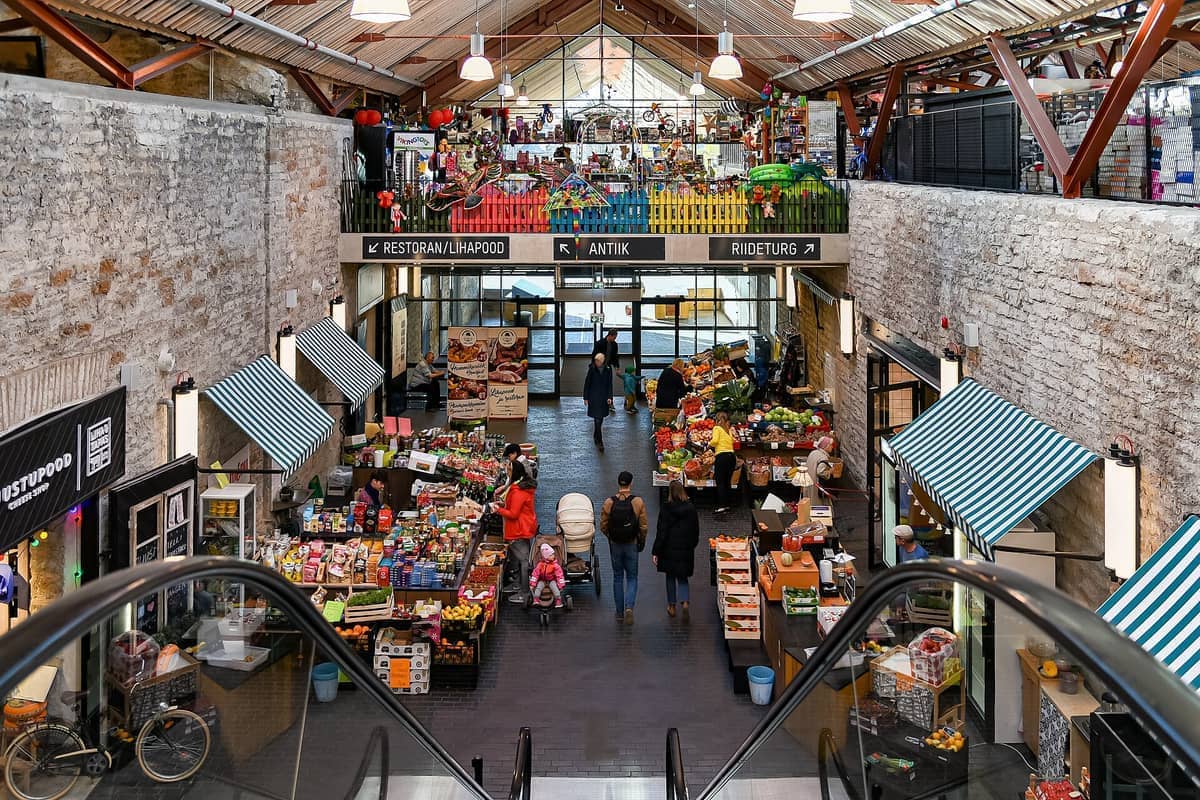Frische Produktstände, Einkäufer und bunte Marktstände im Inneren des Balti Jaam Markts, mit Sichtbacksteinwänden und natürlichem Tageslicht in Tallinn, Estland.