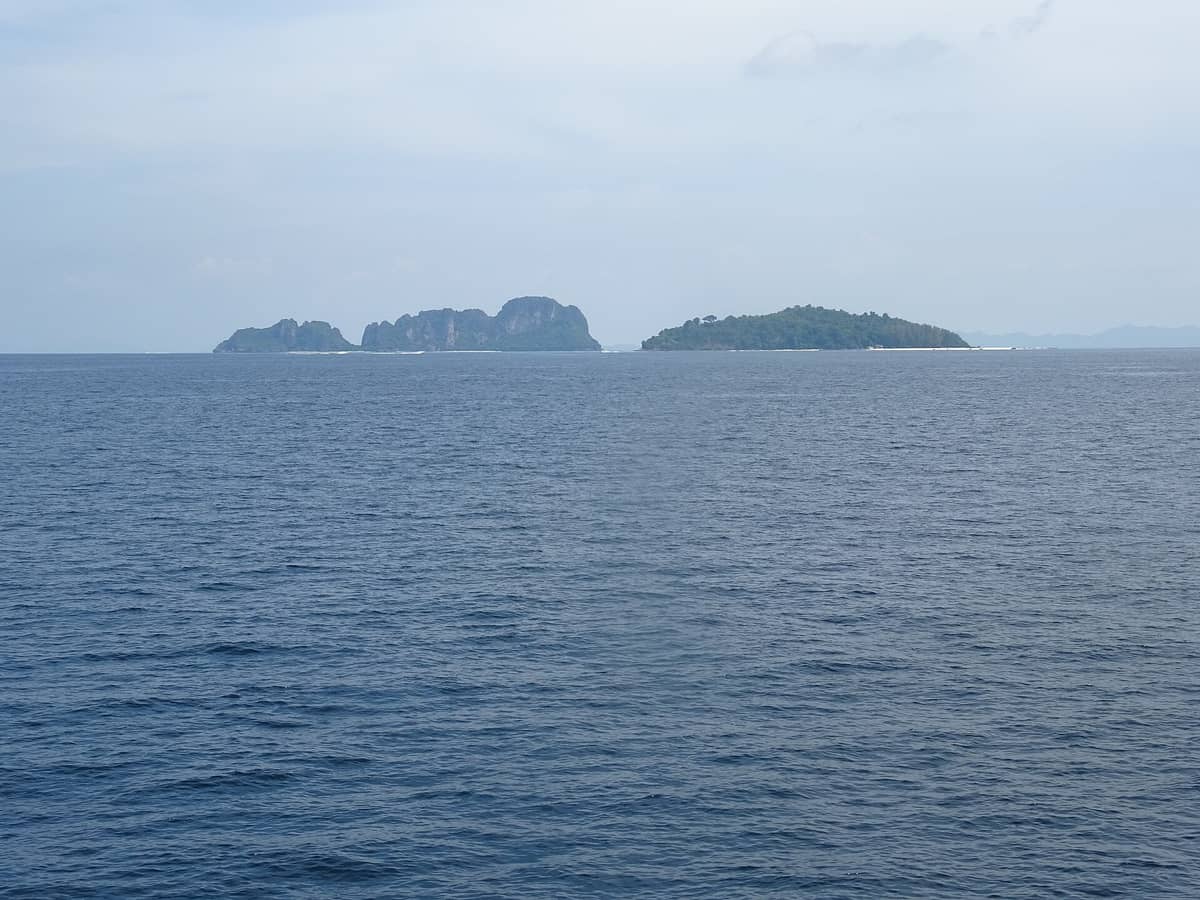 ภาพมุมกว้างจากทะเล ฝั่งขวาเห็นเกาะไม้ไผ่หาดขาวกว้างกับแนวต้นไม้เตี้ย ด้านหลังไกลคือหมู่เกาะพีพี