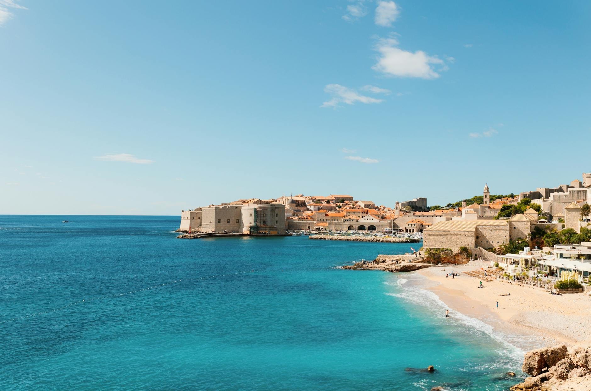 Vista impresionante de la playa Banje con su orilla de guijarros, el agua azul cristalina y las murallas medievales del Casco Antiguo de Dubrovnik al fondo bajo un cielo azul brillante.