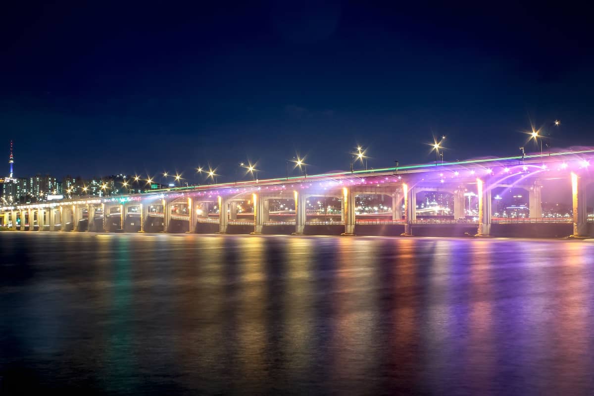 A Ponte Banpo à noite com o show da fonte arco-íris em plena exibição, jatos de água coloridos iluminados por luzes brilhantes ao longo de toda a extensão da ponte sobre o Rio Han.