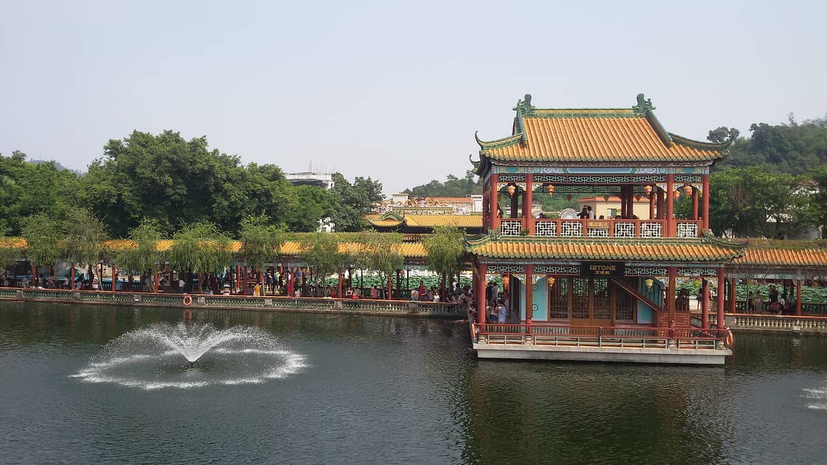 A colorful two-story pavilion with yellow-tiled roof and ornate details sits on a pond with a fountain, surrounded by lush greenery in Baomo Garden.