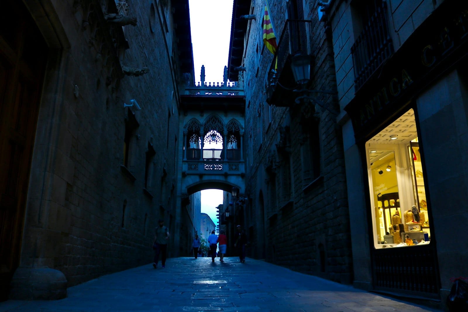 Evening view of Barcelona Gothic Quarter street with iconic bridge, people walking, and inviting shop window lights.