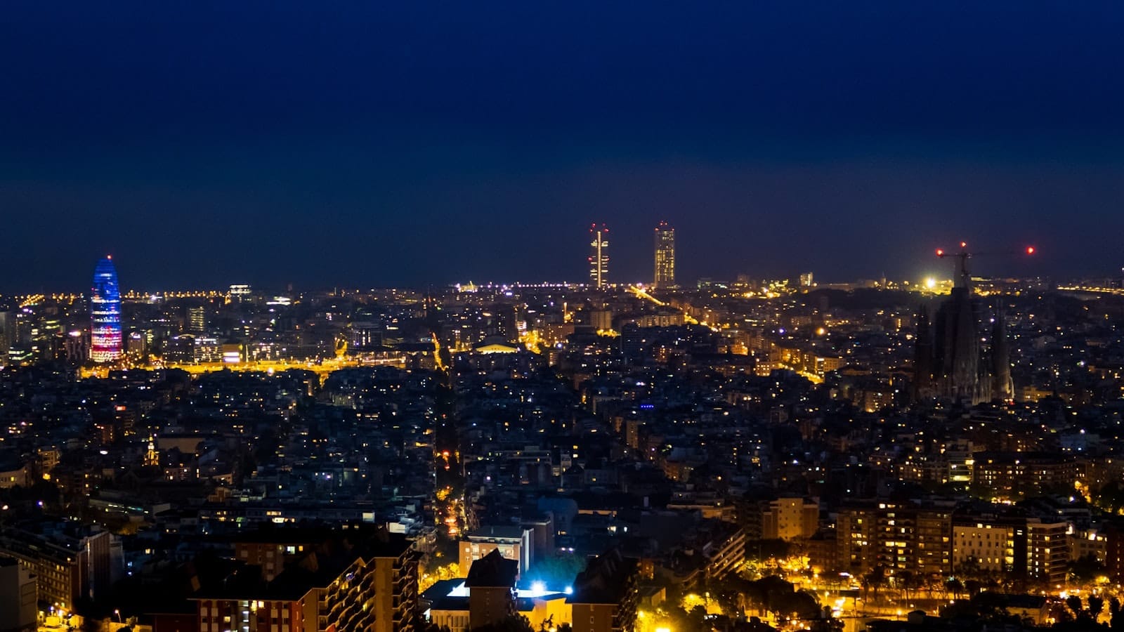 Panoramic nighttime view of Barcelona city skyline with illuminated buildings and deep blue sky, capturing the vibrant urban atmosphere after dark.
