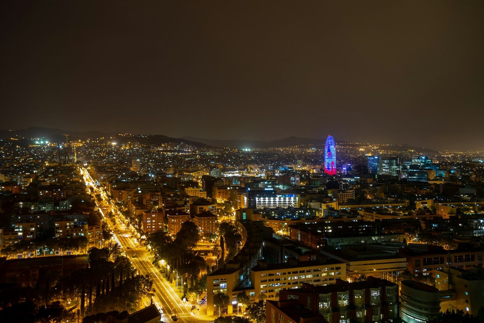 A panoramic nighttime view over Barcelona with city lights stretching toward the horizon, and a prominent illuminated tower in the distance.