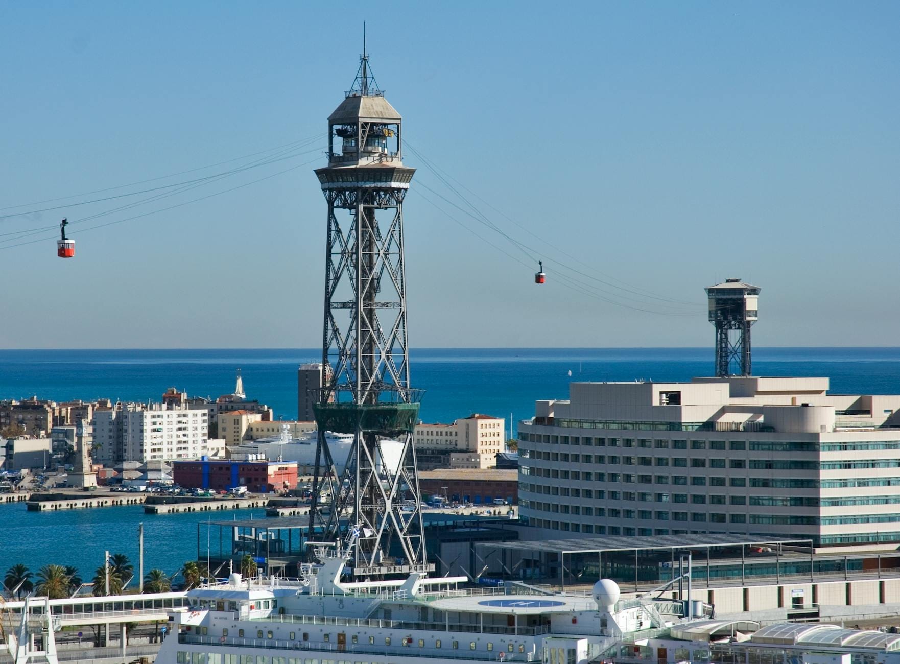Cable cars cross above the Barcelona port area with blue sea, modern buildings, and city skyline visible below.