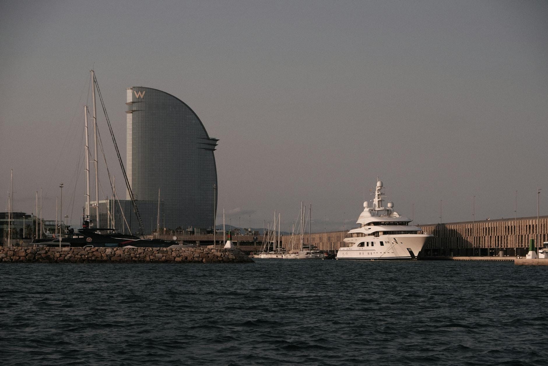 Evening view of the Barcelona waterfront with the iconic W Hotel and yachts moored in the marina, under a dusky sky.