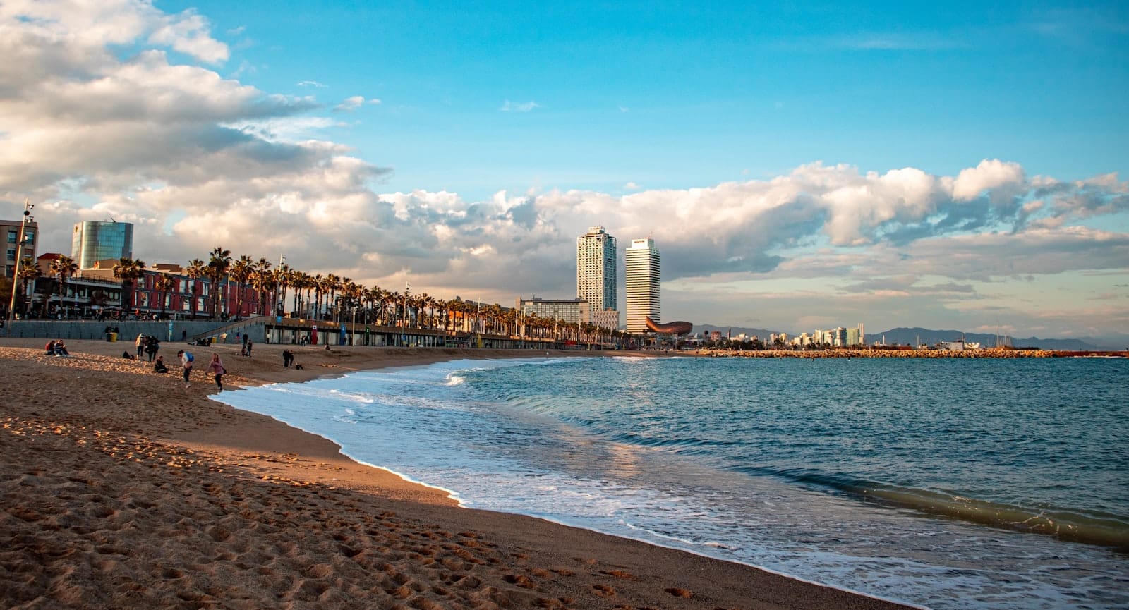 Wide sandy Barcelona beach with gentle waves, city skyline including iconic twin towers and palm trees, all under a vibrant blue sky with fluffy clouds.
