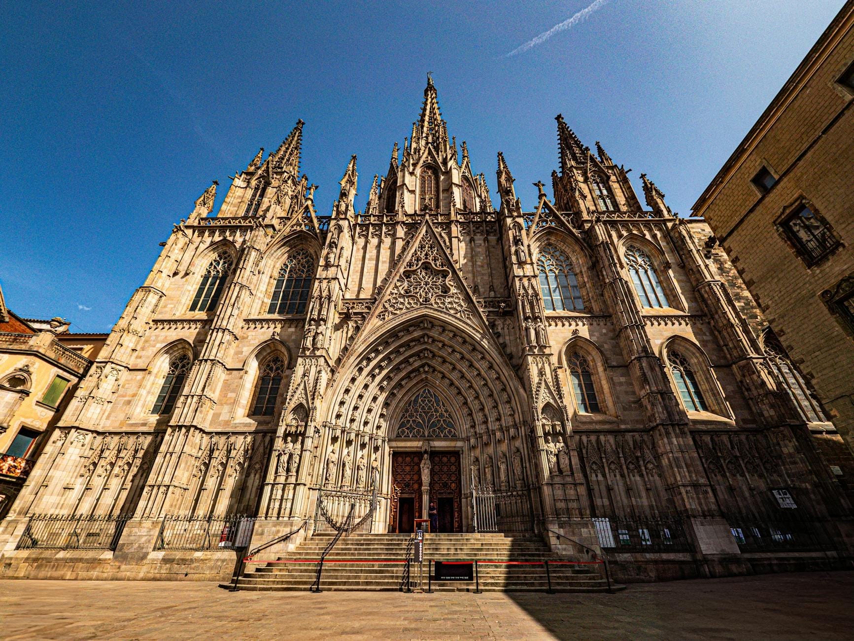 Wide-angle view of Barcelona Cathedral’s ornate Gothic facade under a clear blue sky, with intricate details and grand entrance, inviting visitors to explore its historic architecture.