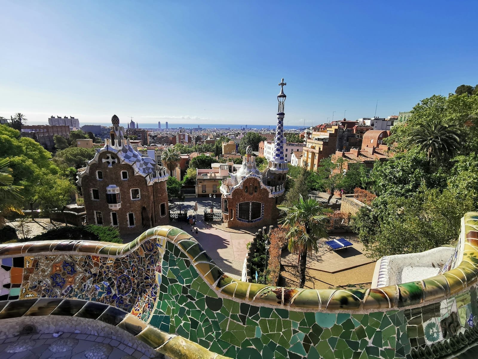 View over Park Güell’s colorful Gaudí mosaic terrace and whimsical gatehouses with Barcelona cityscape in the background under a clear blue sky.