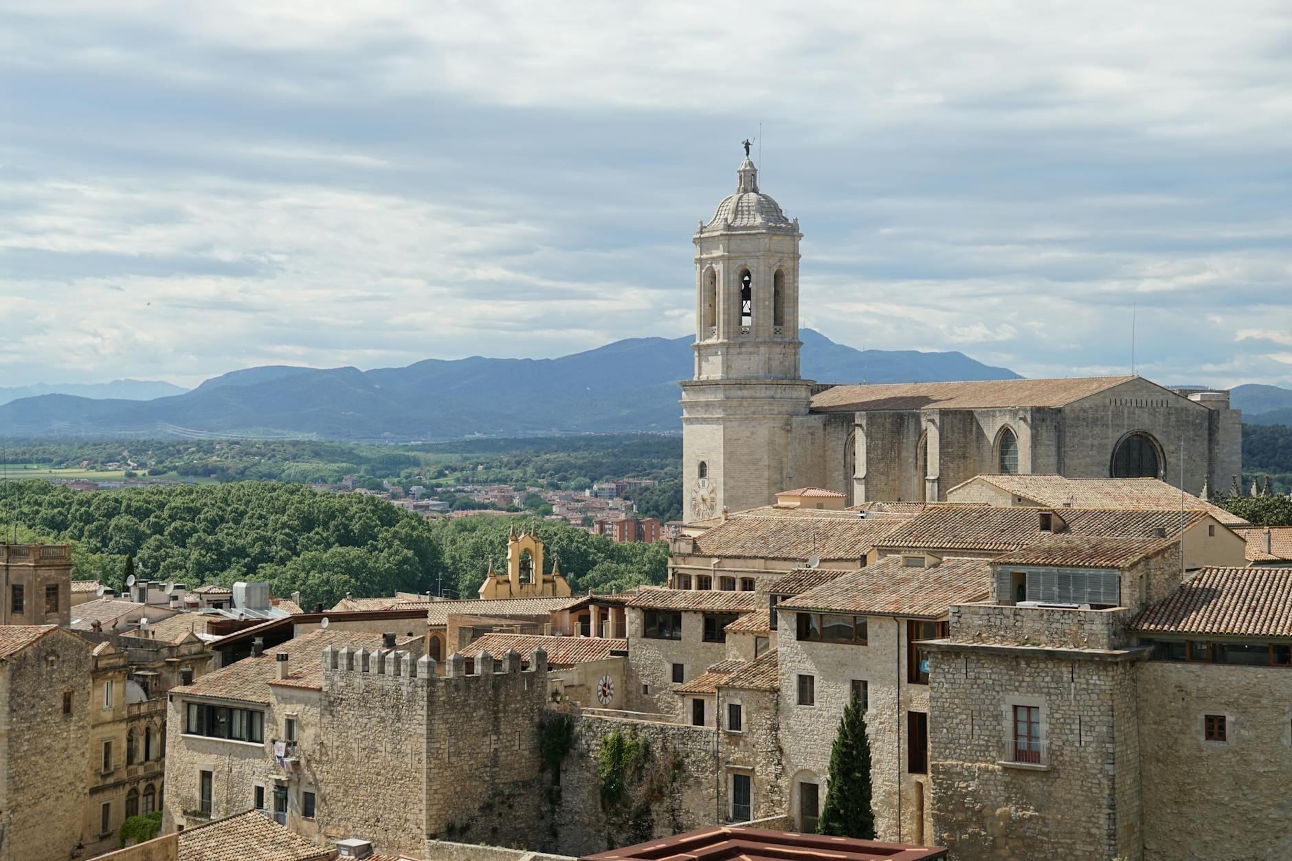 Panoramic view of Girona’s medieval old town with cathedral, stone walls, and distant mountains under a cloudy sky.