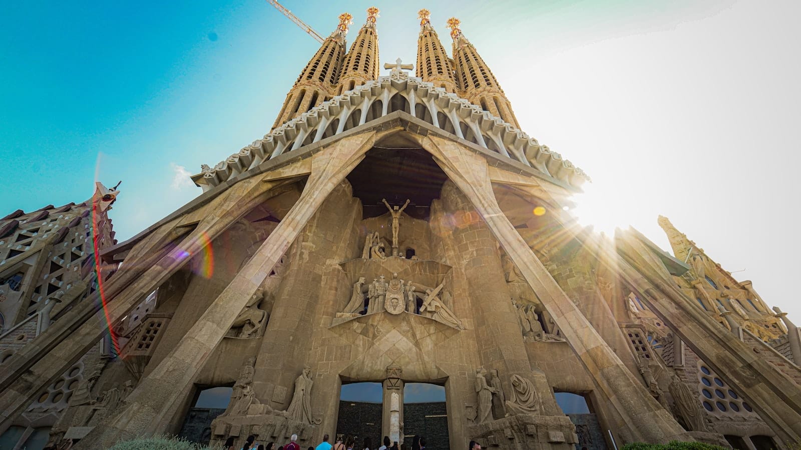 Low-angle view of Sagrada Familia facade with sunlight and clear sky, showing intricate architecture and tall spires under construction.