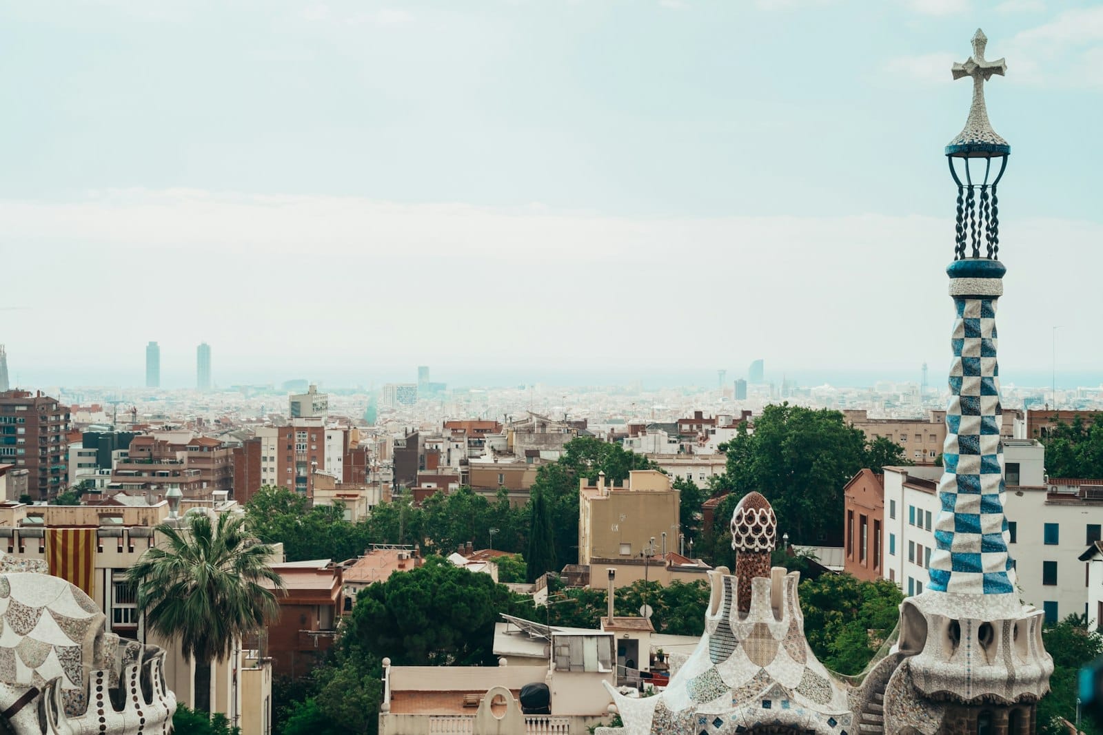 View over Barcelona from Park Güell featuring colorful Gaudí mosaics in the foreground and the cityscape with distant ocean in the background.