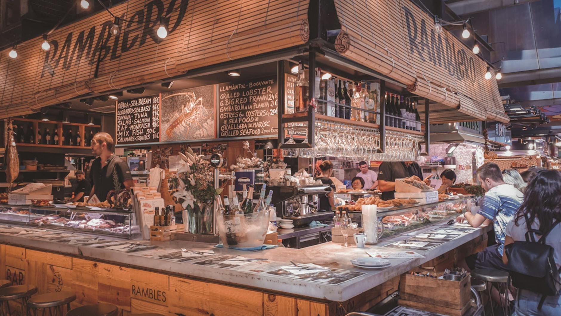 View inside a lively Barcelona market eatery with diners at a bar, handwritten menus, and wine glasses hanging overhead.