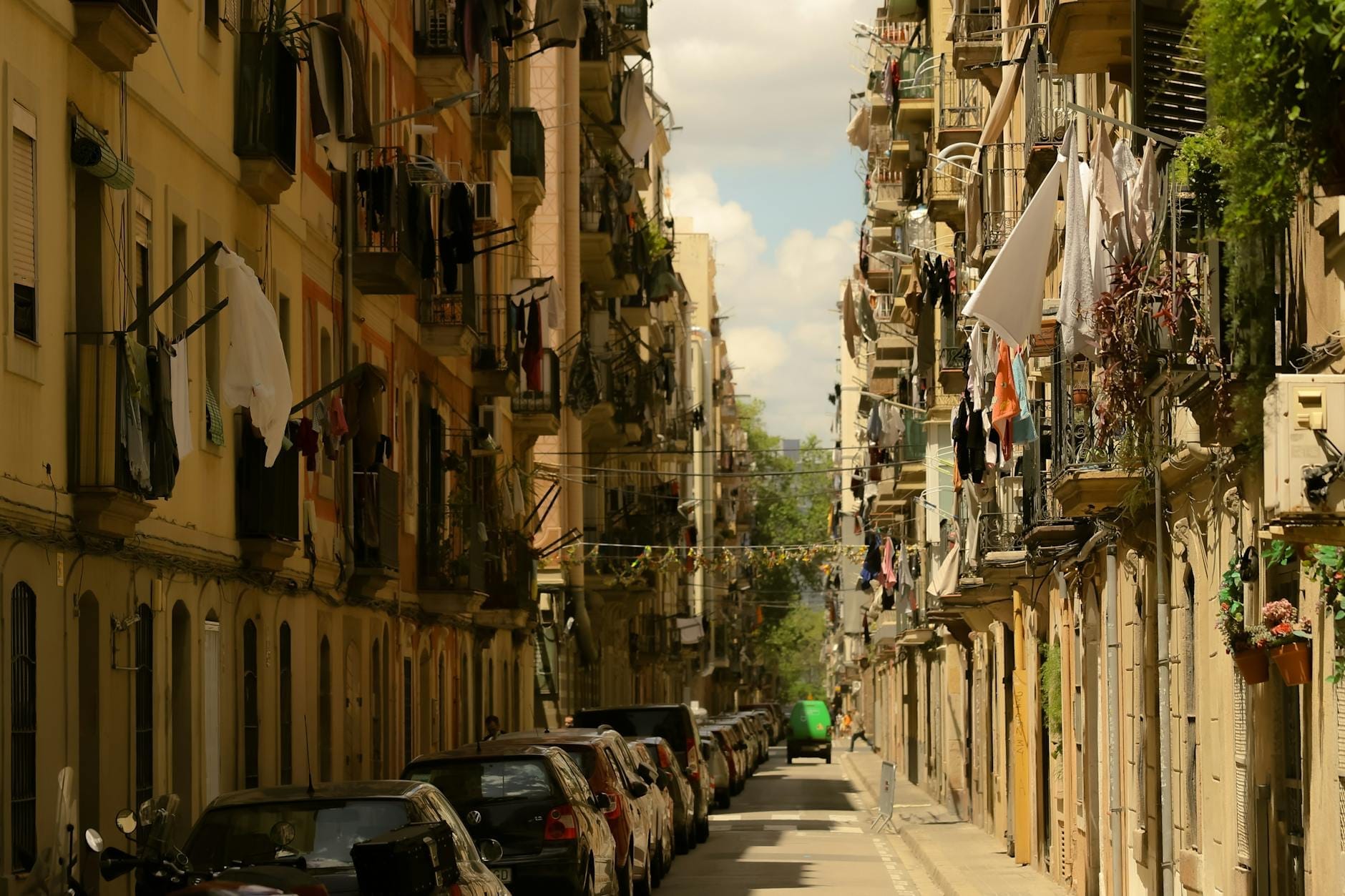 Narrow Barcelona street lined with older apartment buildings, laundry hanging from balconies, and parked cars along both sides.