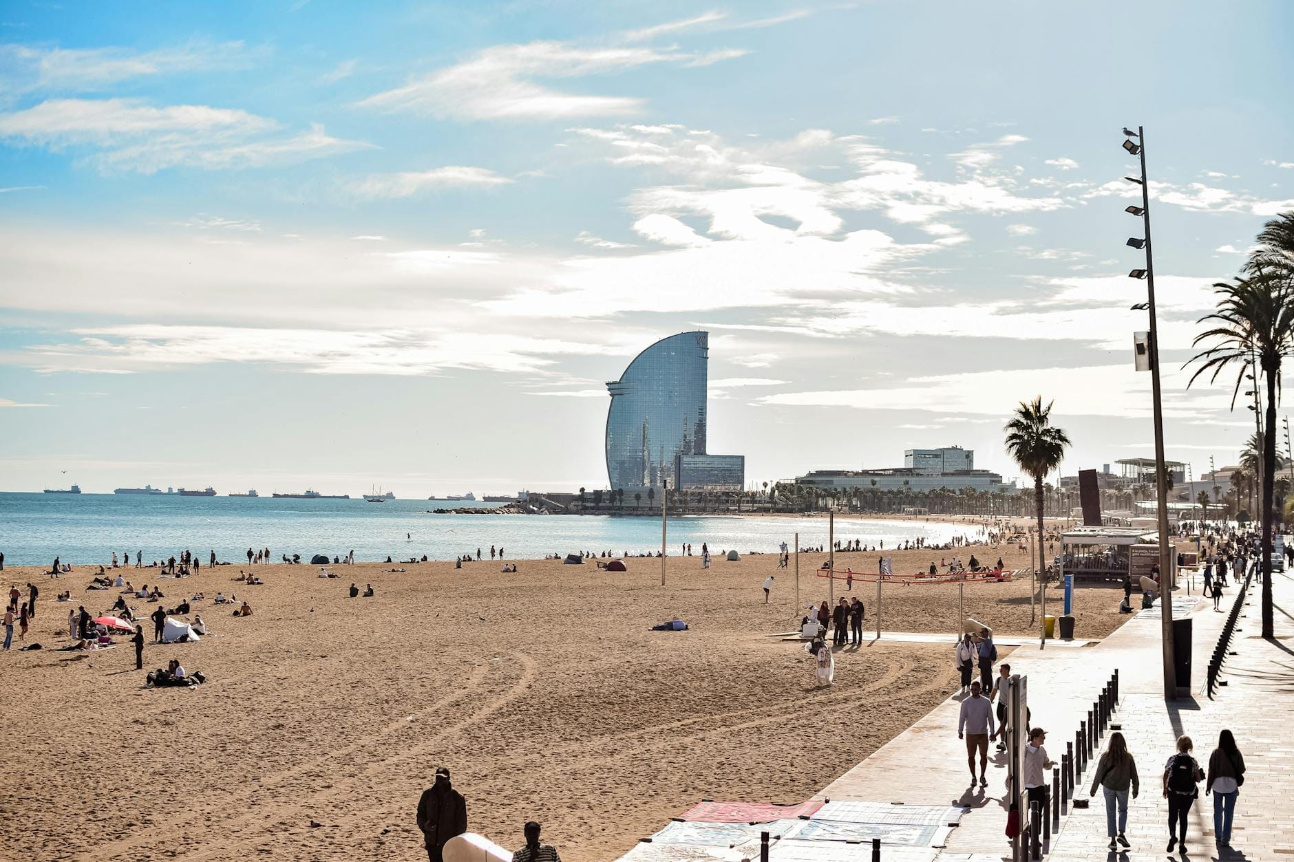 Wide view of Barcelona's Barceloneta beach with the sea, promenade, scattered visitors, and iconic W Hotel in the background under blue sky.