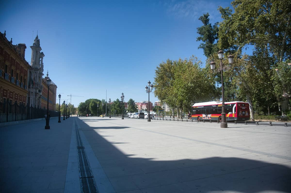 รถบัสสีแดงและขาวจอดอยู่บนถนนกว้างที่เรียงรายด้วยต้นไม้ในเมือง Seville พร้อมอาคารประวัติศาสตร์และท้องฟ้าสีฟ้าใส