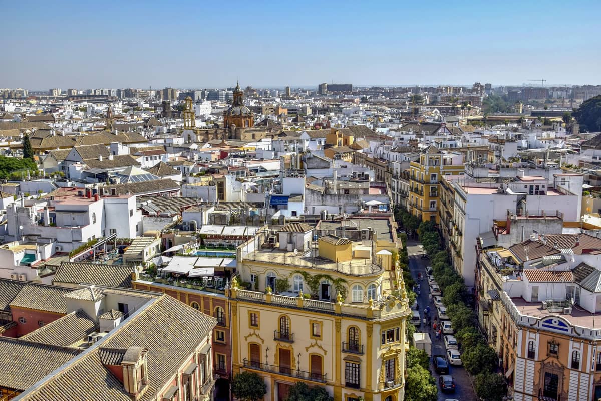 ภาพถ่ายทางอากาศมุมกว้างของย่านใจกลางเมือง Seville พร้อมอาคารประวัติศาสตร์ ถนนที่เรียงรายด้วยต้นไม้ และท้องฟ้าสีฟ้าใสเหนือขึ้นไป