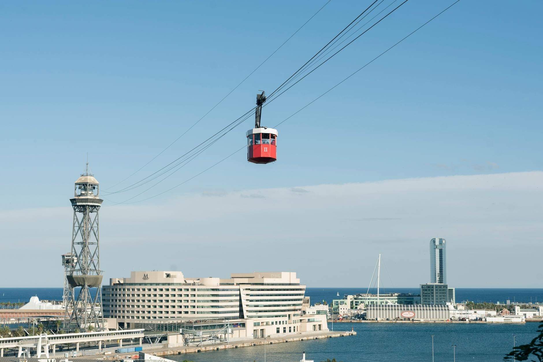 A red Montjuïc cable car travels above Barcelona’s waterfront with iconic towers, blue sea, and modern city buildings under a clear sky.