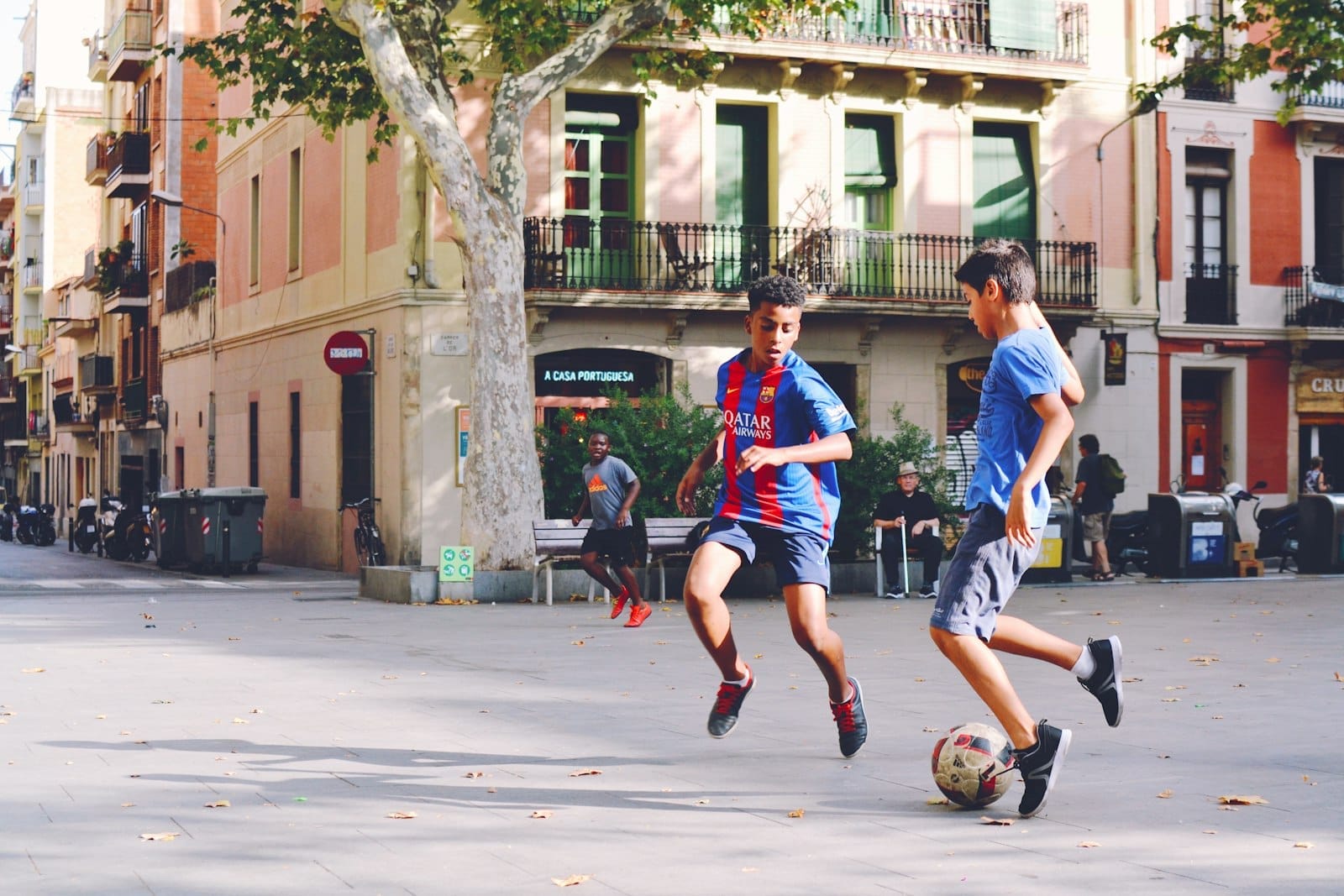Two children playing soccer in a sunny Barcelona plaza surrounded by colorful buildings and trees, capturing playful energy for family travel.