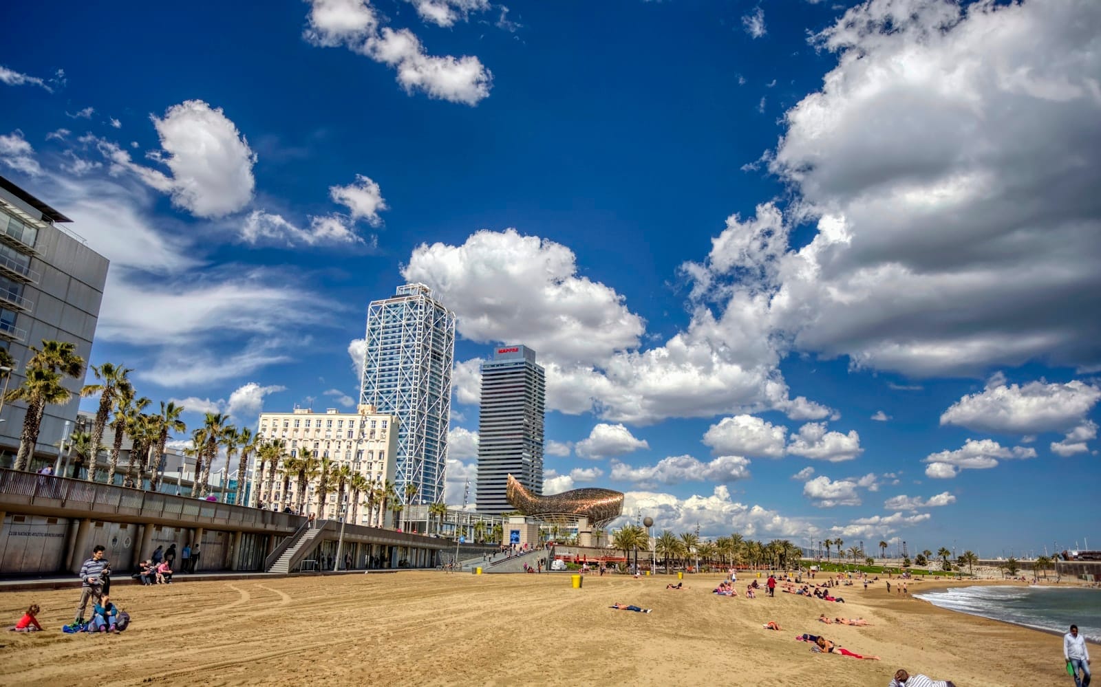 Barceloneta beach with people relaxing on the sand, iconic waterfront towers, palm trees, and a vibrant sky filled with dramatic clouds.