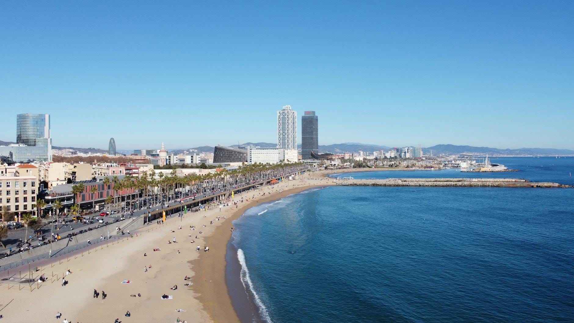 Aerial view of Barceloneta Beach in Barcelona with city skyline, palm-lined promenade, golden sand, and blue Mediterranean sea on a sunny day.