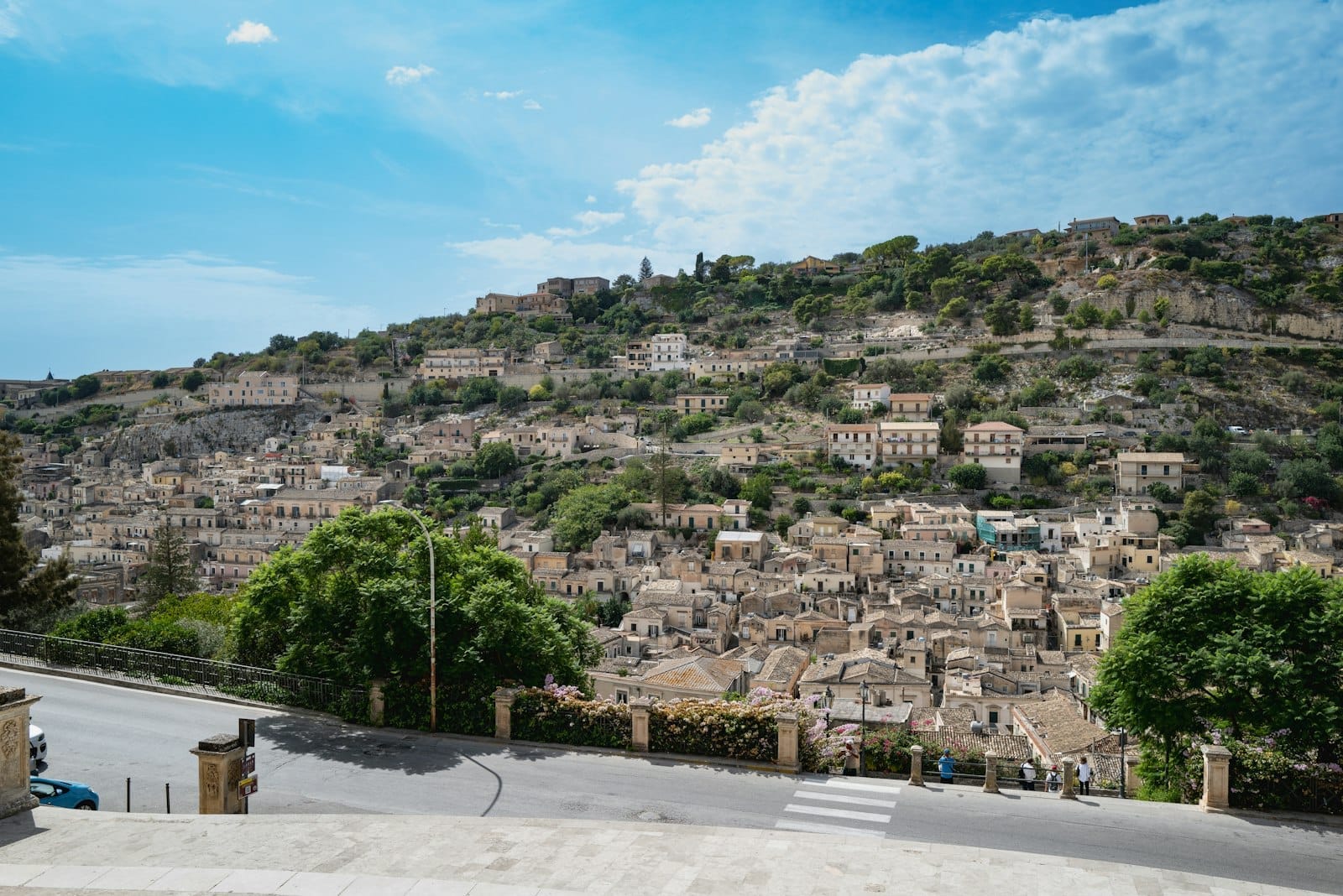 Panoramablick auf die barocke Hügelstadt Modica in Sizilien, mit dicht gedrängten Steingebäuden auf grünen Hängen unter einem hellen Himmel.