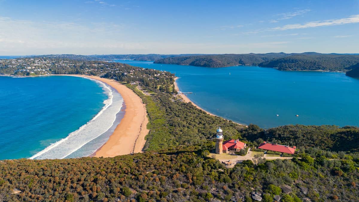 Vista aérea del faro de Barrenjoey sobre el banco de arena de Palm Beach, con Pittwater y el océano Pacífico a cada lado bajo un cielo azul brillante.