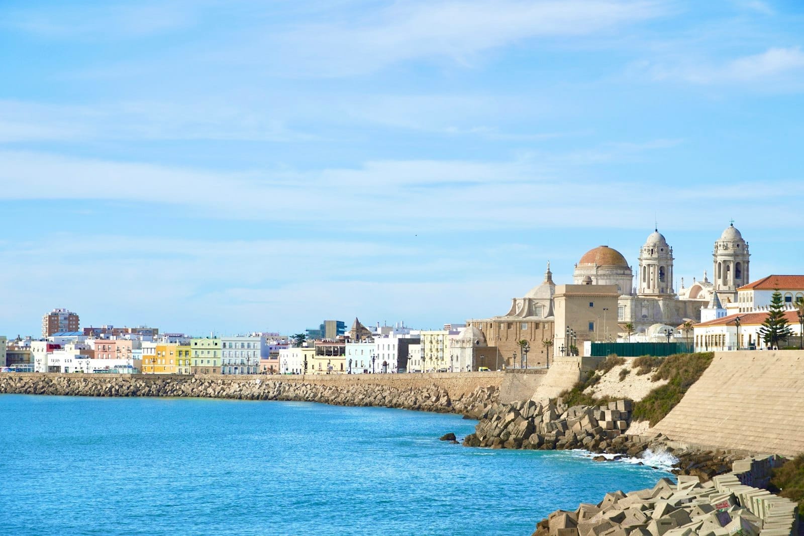 Vista panoramica del centro storico di Cádiz con la Cattedrale e le antiche mura sul lungomare che si affacciano sul mare azzurro sotto un cielo luminoso.