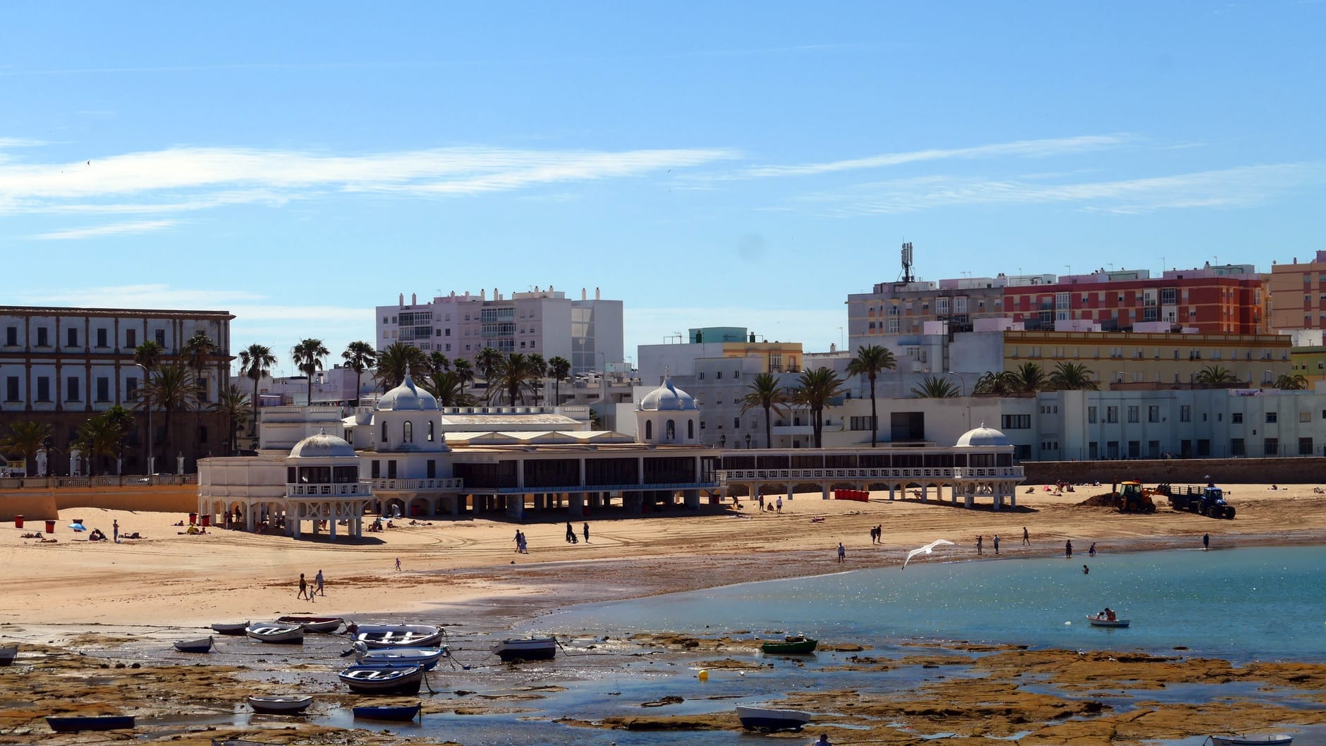 Vista sul lungomare del Barrio de La Viña a Cádiz, con edifici imbiancati, piccole imbarcazioni sulla sabbia e palme sotto cieli azzurri e sereni.