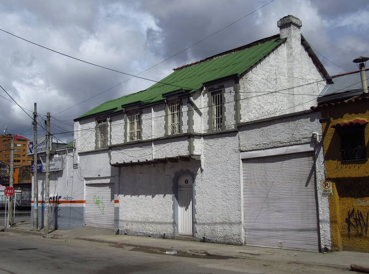 Street view of a modest residential and commercial building with a green roof and white facade in Barrio San Felipe, Bogotá under cloudy skies.