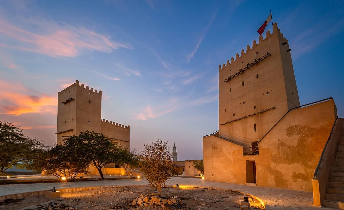 Barzan Towers in der Abenddämmerung, zwei historische Wachtürme mit Zinnen und der Flagge Katars, umgeben von Bäumen und sanfter Wegbeleuchtung.
