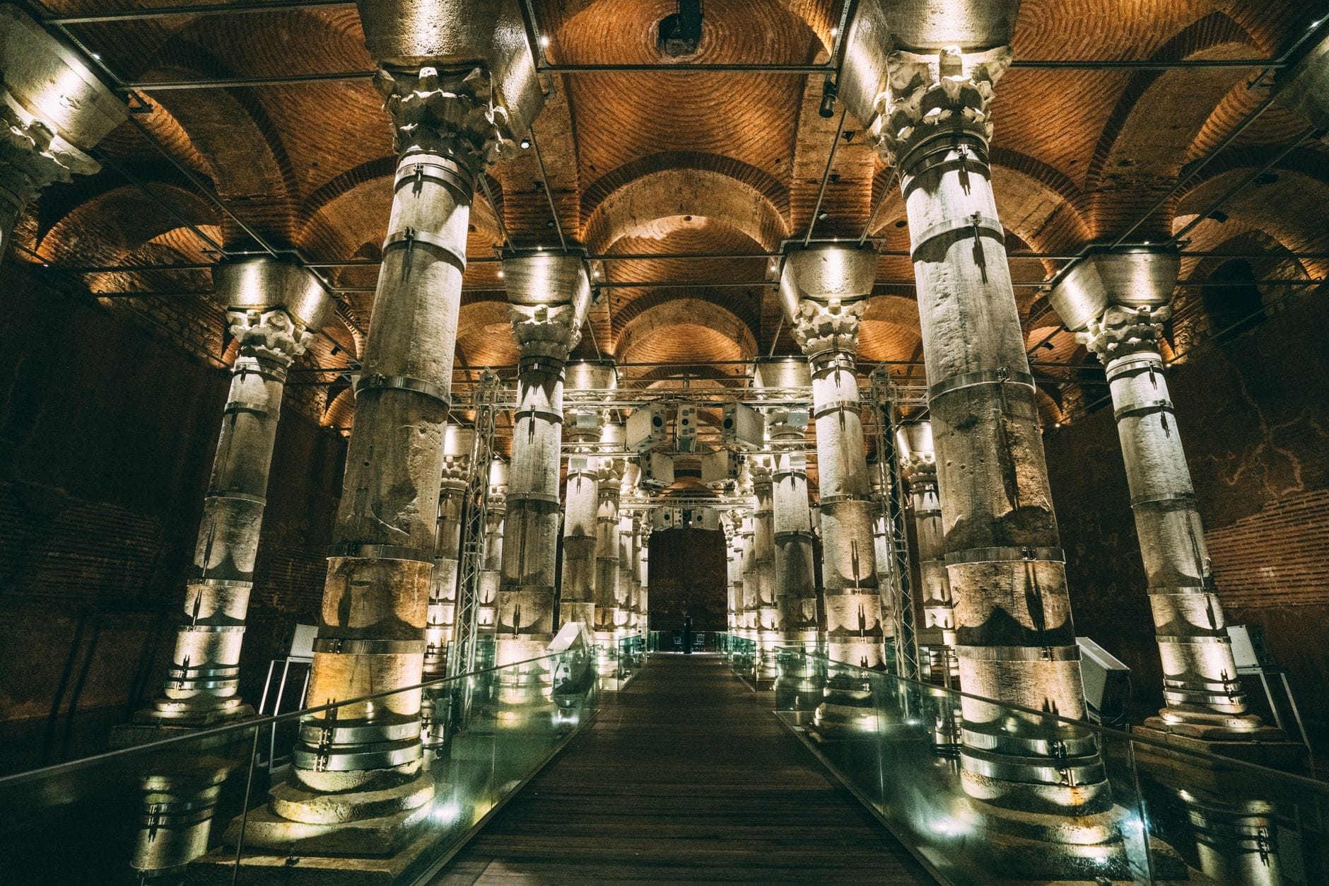 Vista ampia dell'interno della Cisterna Basilica con antiche colonne illuminate, soffitti a volta in mattoni e un camminamento centrale che si riflette sull'acqua.