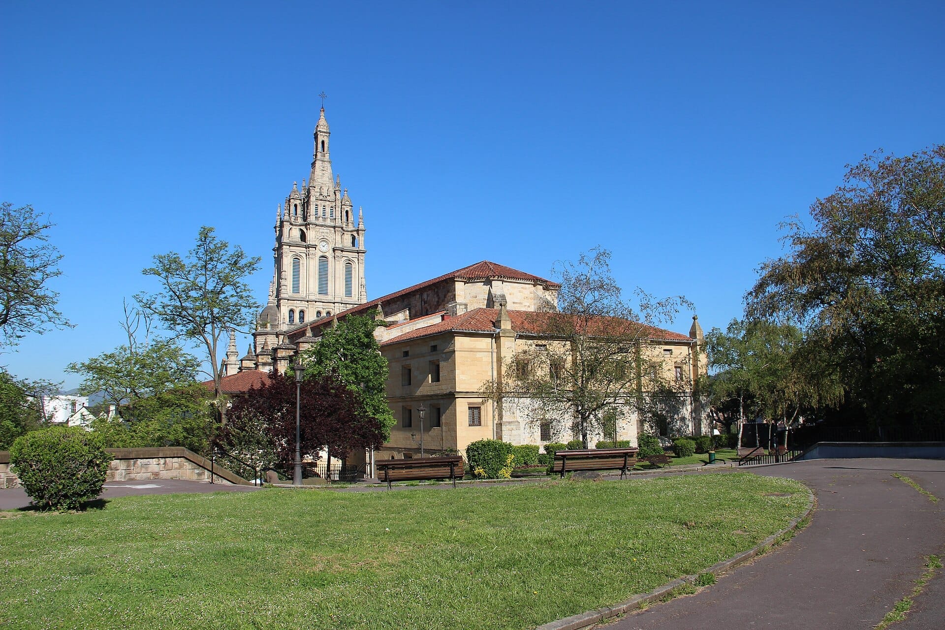 The Basílica de Begoña in Bilbao, seen from a grassy park area with benches, surrounded by trees and clear blue sky.