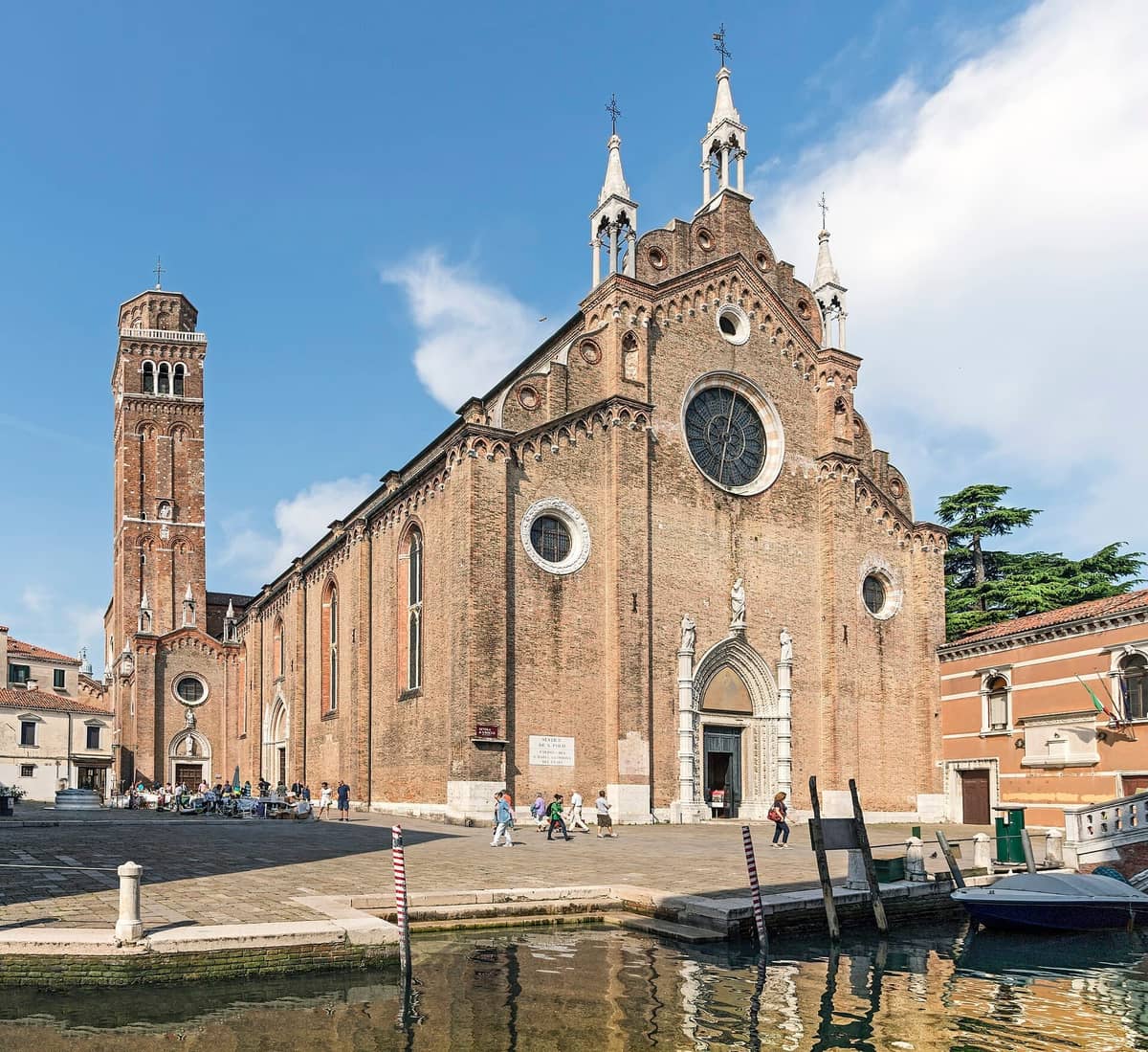 Außenansicht der Basilica dei Frari in Venedig mit ihrer hohen Backsteinfassade, dem Rosettenfenster und dem kanalzugewandten Eingang unter einem klaren blauen Himmel.