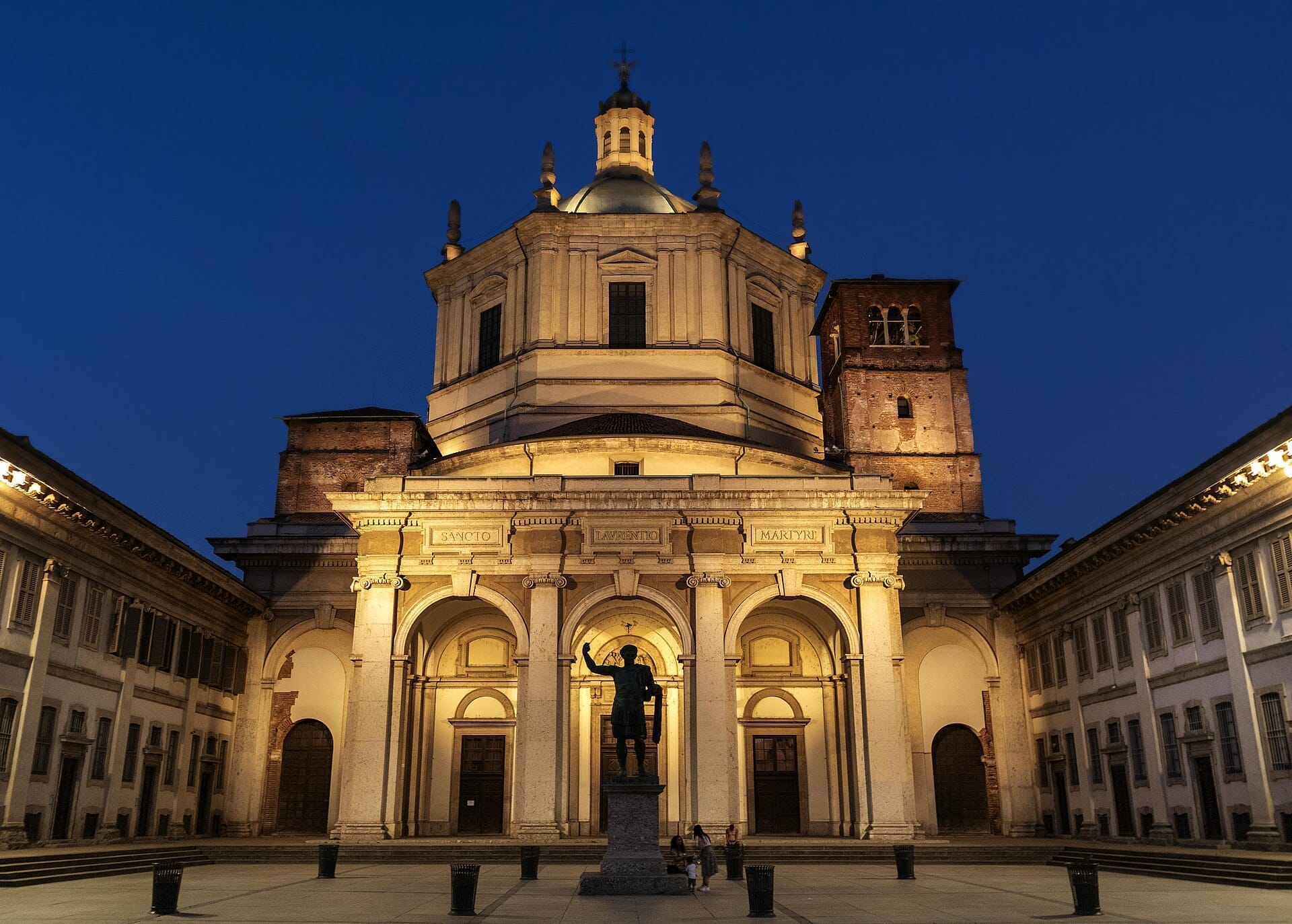 Basilica di San Lorenzo Maggiore illuminata al tramonto, con colonne romane, la cupola centrale e una statua in primo piano.