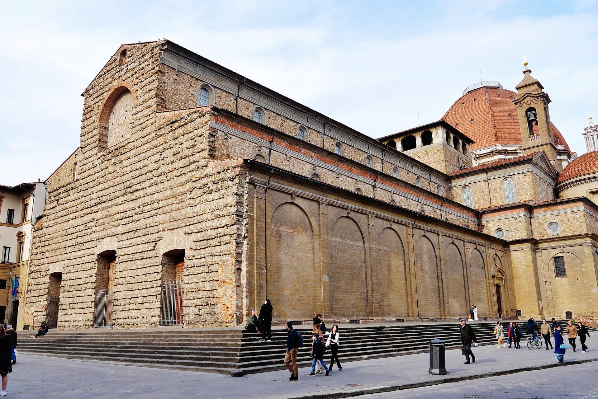 มุมมองเฉียงของฟาซาดหินที่ยังสร้างไม่เสร็จและโดมของ Basilica di San Lorenzo ในฟลอเรนซ์ มีผู้คนเดินเล่นท่ามกลางแสงแดดในจัตุรัสหน้าโบสถ์