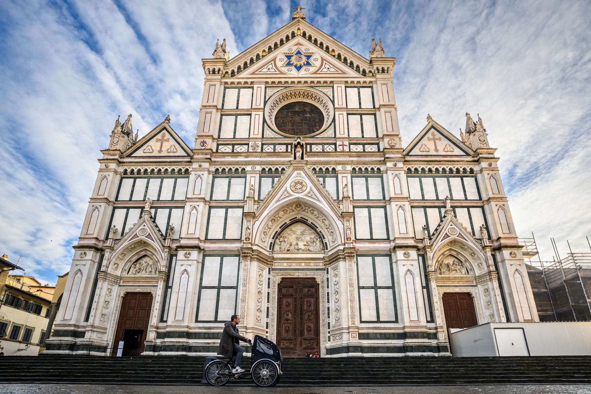 วิวกว้างที่เห็นด้านหน้าหินอ่อนของ Basilica di Santa Croce ในฟลอเรนซ์ยามท้องฟ้าแจ่มใส มีชายคนหนึ่งเข็นจักรยานผ่านหน้าวิหาร