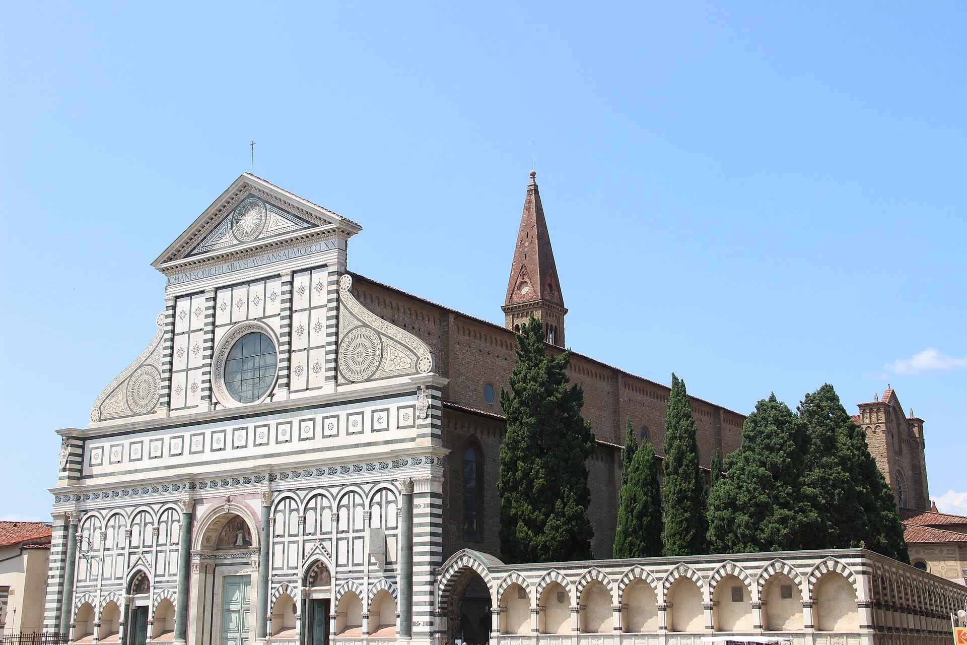 มุมมองหน้าจั่วหินอ่อนลายเรขาคณิตของ Basilica di Santa Maria Novella หอระฆังสูงและต้นไม้รอบ ๆ ใต้ท้องฟ้าสีฟ้าสดใส