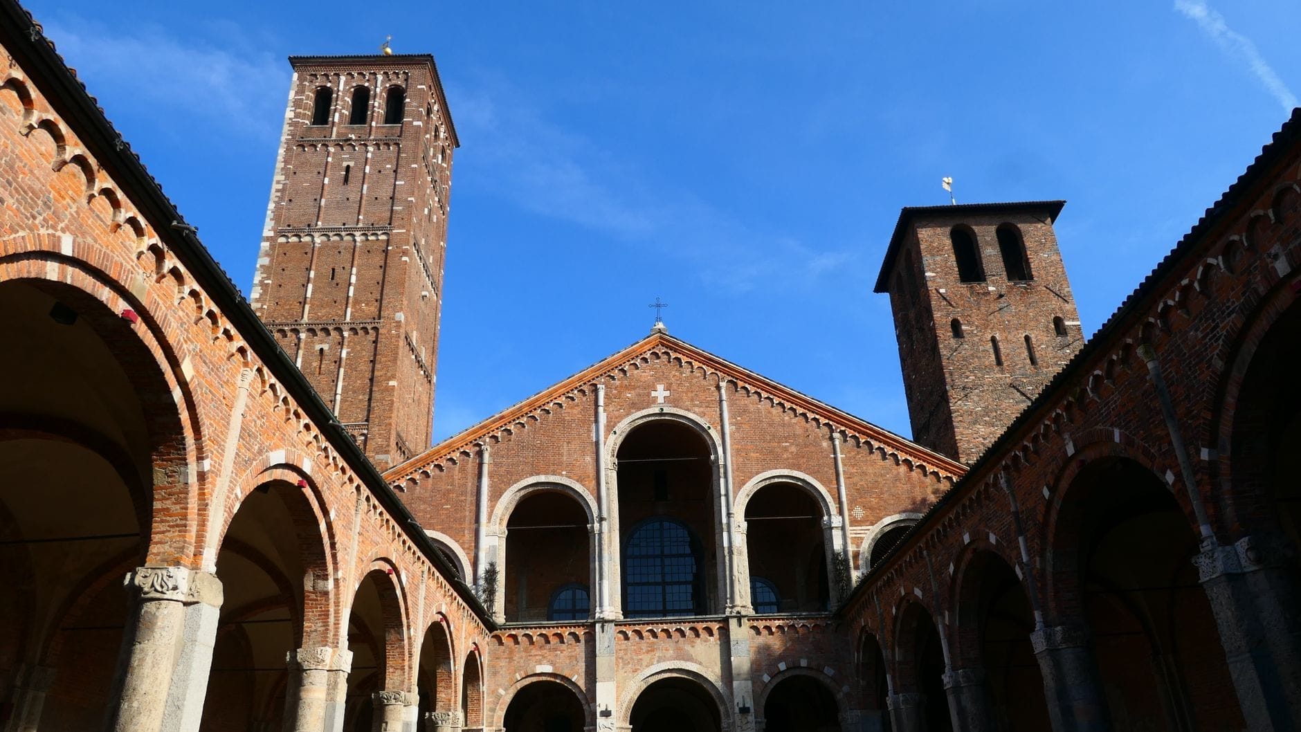 Vista del cortile e della facciata della Basilica di Sant'Ambrogio con le due torri romaniche e il portale ad arco, sotto un cielo azzurro limpido a Milano.