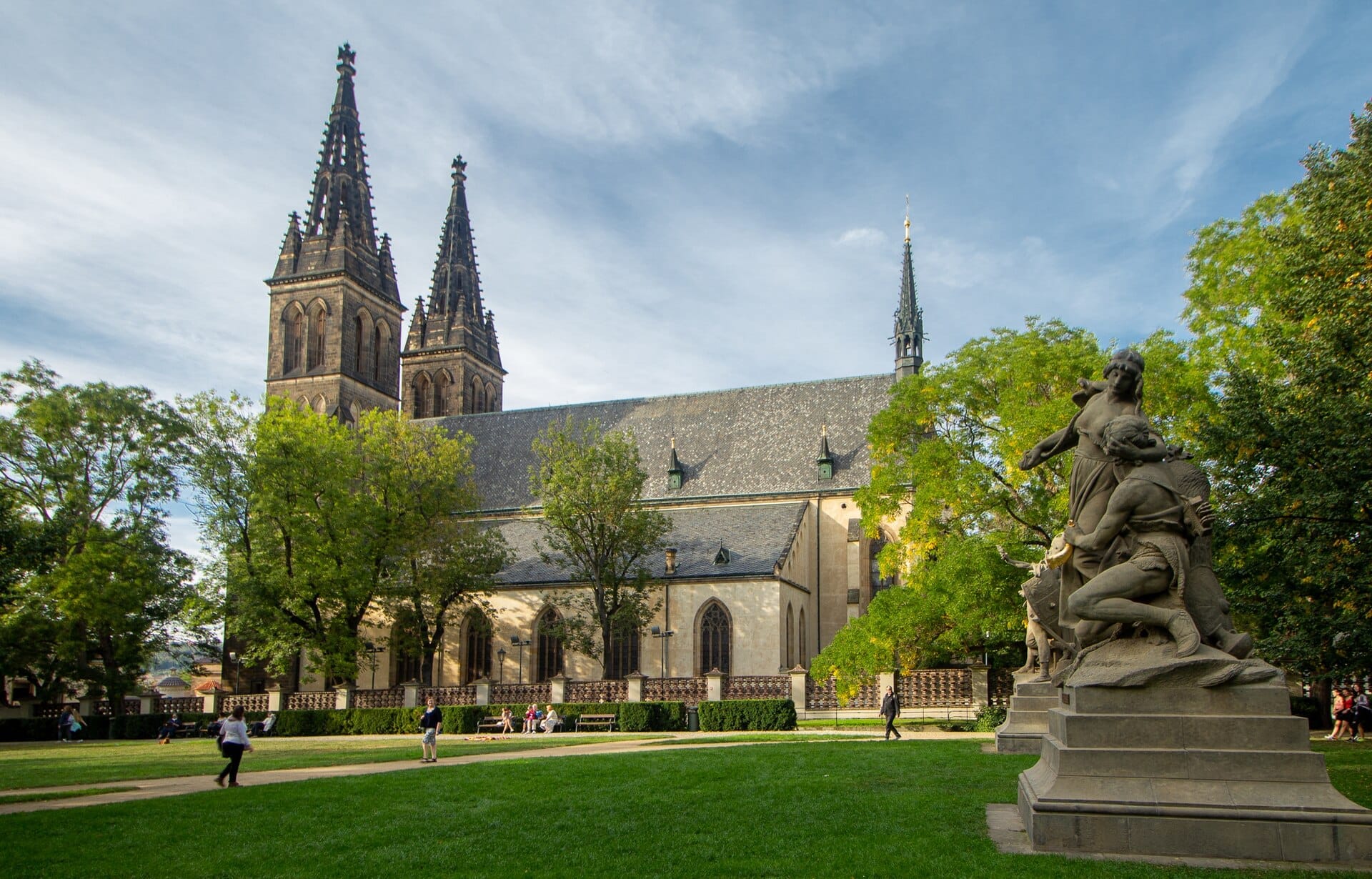 Die Basilika St. Peter und Paul in Vyšehrad in Prag, mit zwei neugotischen Türmen, grünen Bäumen und Statuen unter einem blauen Himmel.