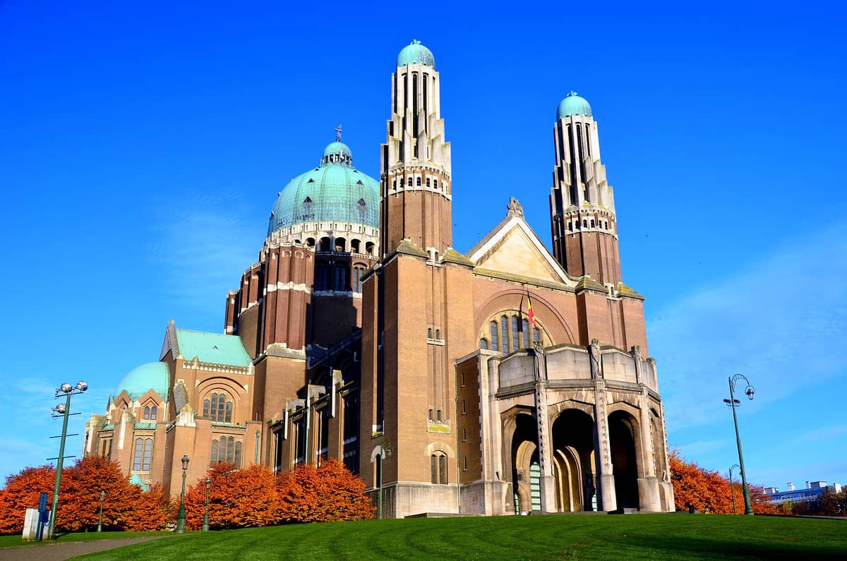 Vista exterior da Basílica do Sagrado Coração em Koekelberg, Bruxelas, com sua distinta cúpula verde Art Déco, duas torres e fachada de tijolos marrons sob um céu azul intenso.