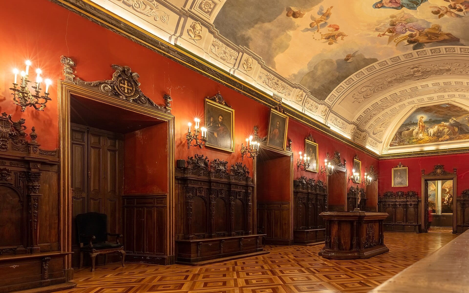 Sala interna ornata della Basílica de San Francisco el Grande con pareti rosse, panche in legno, applique stile candela, ritratti e soffitto a volta dipinto.
