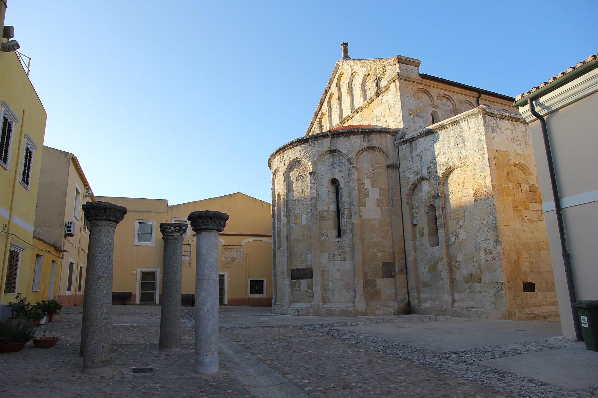 Steinsäulen und die runde Apsis der Basilica di San Gavino in einem sonnigen Innenhof, umgeben von pastellgelben Gebäuden in Porto Torres.