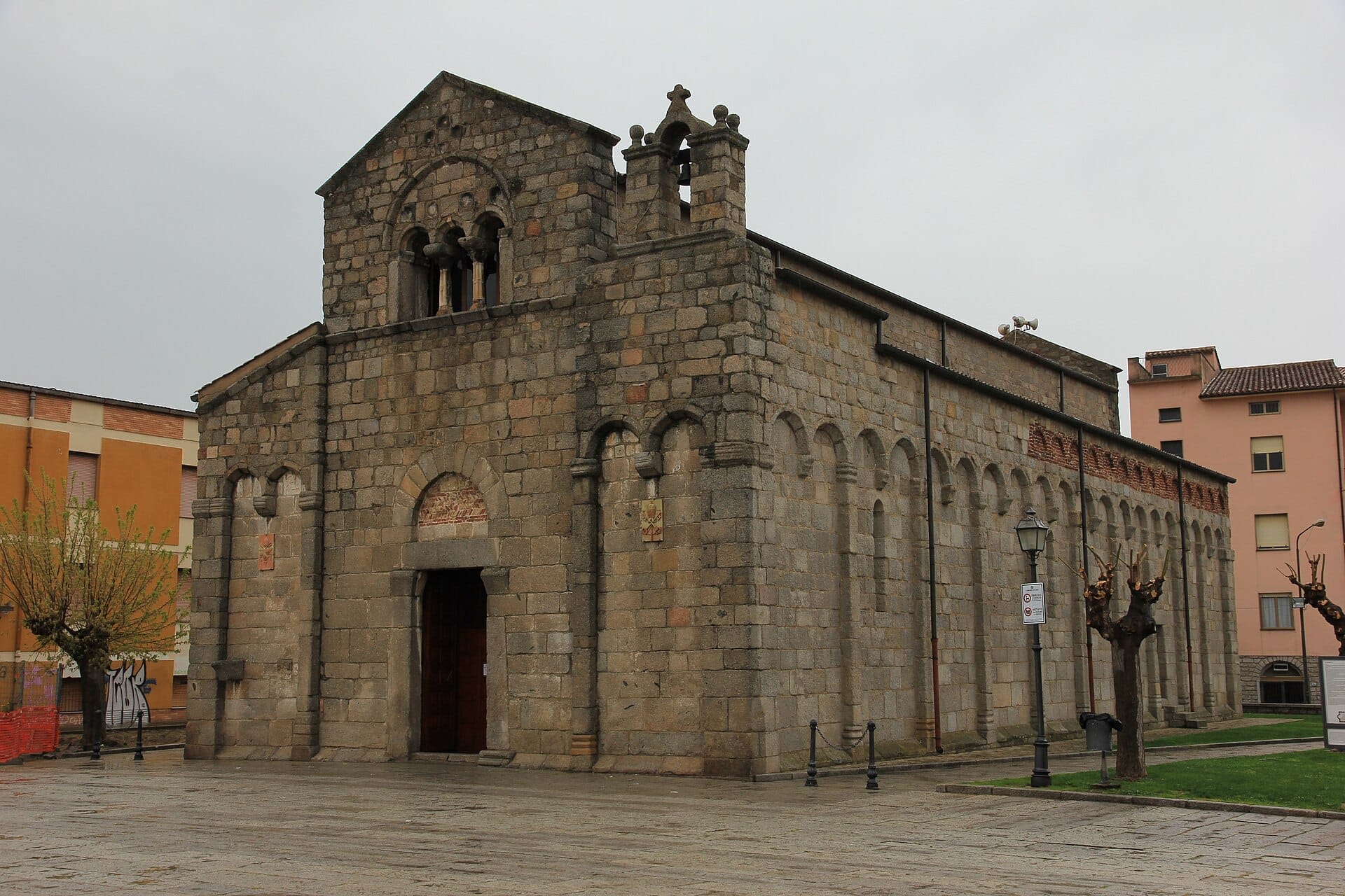 Frontansicht der steinernen Fassade und der romanischen Bögen der Basilica di San Simplicio, umgeben von einem gepflasterten Platz und gestutzten Bäumen.