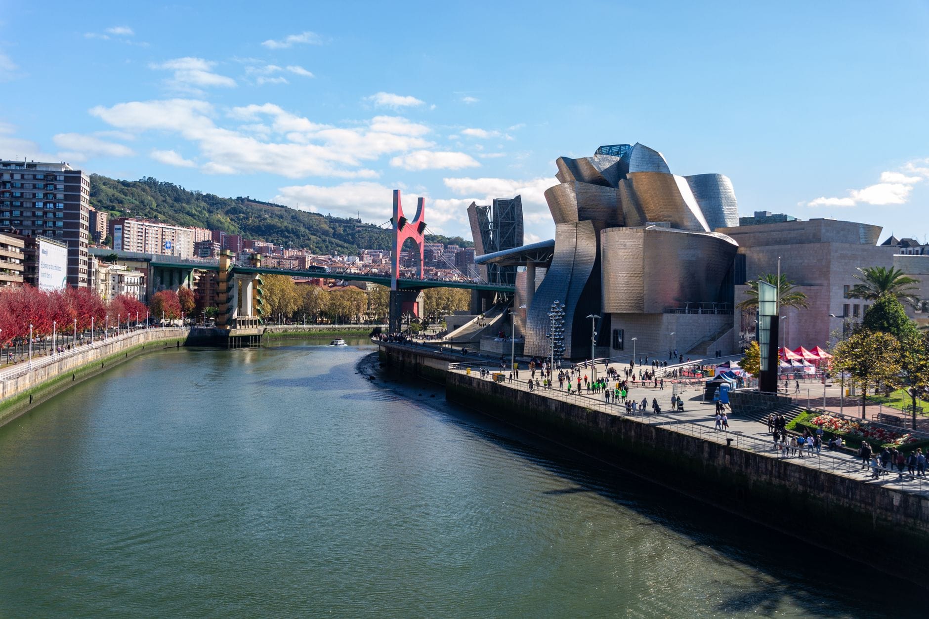 Tageslichtblick auf die Uferpromenade des Guggenheim Museum Bilbao mit seinen Titankurven, der roten Brücke und Stadtgebäuden unter blauem Himmel mit Menschen im Freien.