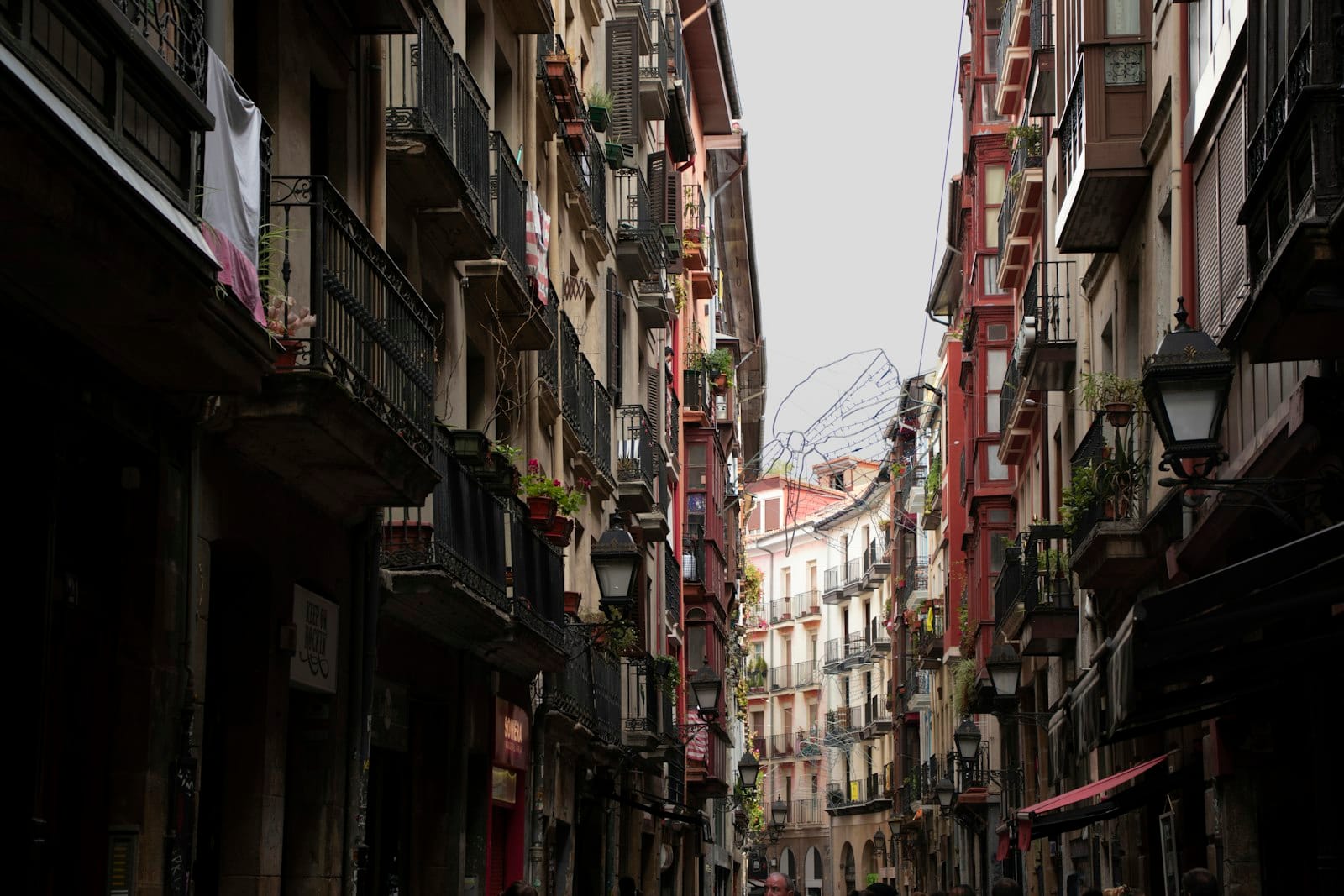 Hohe, schmale historische Gebäude mit Balkonen säumen eine geschwungene Fußgängerzone im Casco Viejo von Bilbao unter einem bedeckten Himmel.