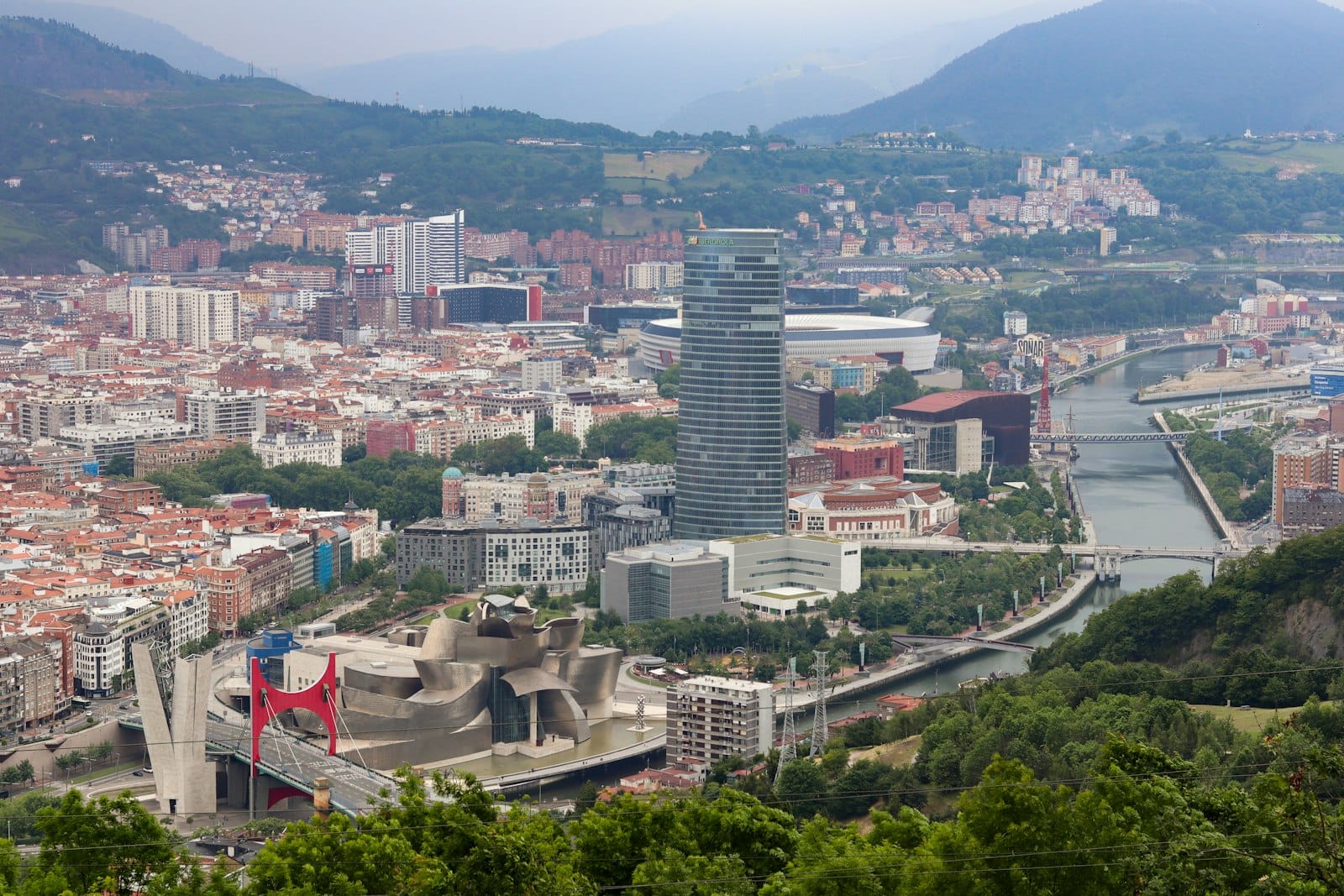 Weitläufige Erhöhungsansicht über Bilbao mit grünen Hügeln, der Stadtsilhouette und Wahrzeichen wie dem Guggenheim Museum und dem Iberdrola Tower.