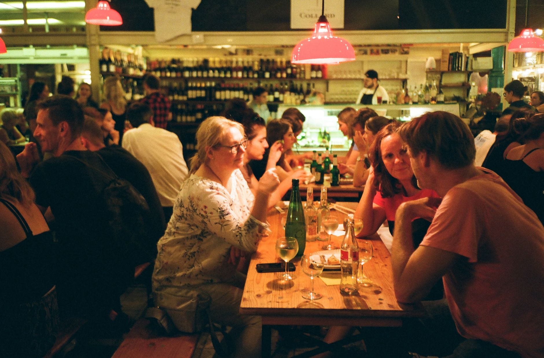 Gruppi di persone che gustano cibo e bevande ai tavoli comuni in un vivace locale interno, con bottiglie e un bar affollato sullo sfondo